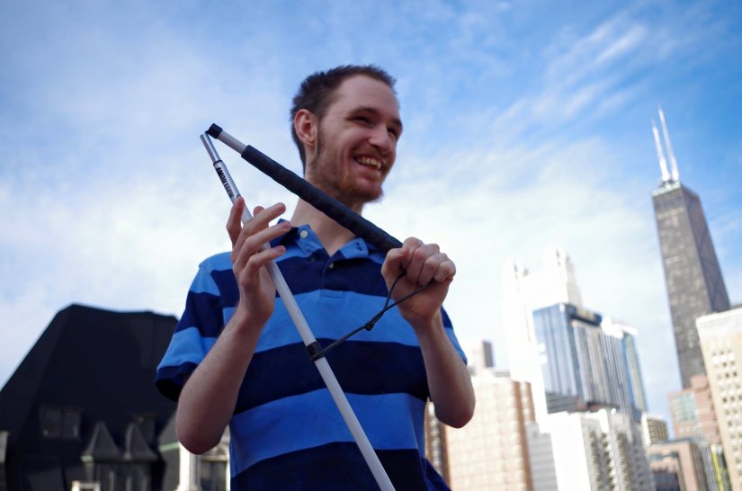 Image of a young man smiling against the backdrop of a city. He is holding a white cane and wearing a striped black and blue shirt. 